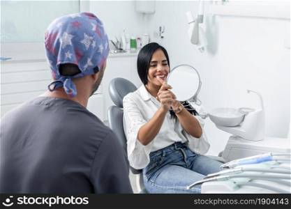 Female client smiling while looking to a mirror in a dental clinic next to a dentist. Cient smiling and looking to a mirror in a dental clinic