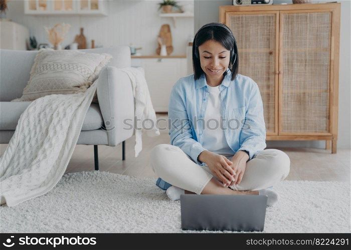 Female coach in headset conducts online lesson on laptop by video call sitting on the floor at home. Friendly smiling young woman consulting client remotely on computer. Distant education, remote job.. Female coach in headset conducts online lesson on laptop by video call sitting on the floor at home