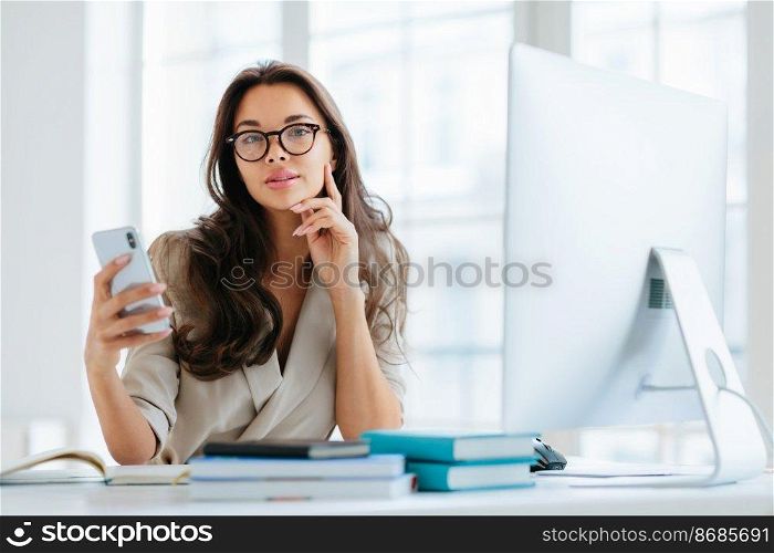 Female company office worker in formal wear uses modern gadget for work, poses at workplace near monitor, plans working schedule, browses information online, poses against window. Working time
