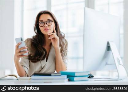 Female company office worker in formal wear uses modern gadget for work, poses at workplace near monitor, plans working schedule, browses information online, poses against window. Working time
