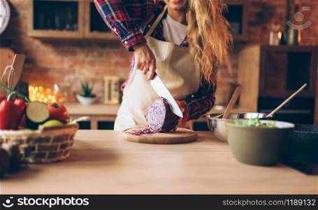 Female cook in apron cutting fresh vegetables, kitchen interior on background. Housewife with knife in hands making healthy vegetarian food, salad preparation. Female cook in apron cutting fresh vegetables