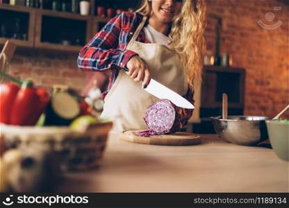 Female cook in apron cutting fresh vegetables, kitchen interior on background. Housewife with knife in hands making healthy vegetarian food, salad preparation. Female cook in apron cutting fresh vegetables
