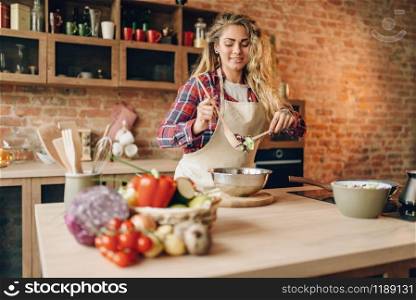 Female cook in apron prepares fresh salad, kitchen interior on background. Housewife making healthy vegetarian food, vegetables preparation. Female cook in apron prepares fresh salad