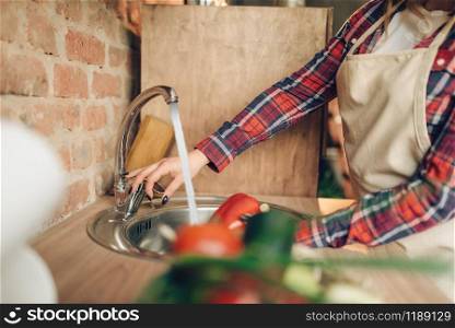 Female cook in apron washes fresh vegetables, kitchen interior on background. Housewife making healthy vegetarian food, salad preparation. Female cook in apron washes fresh vegetables