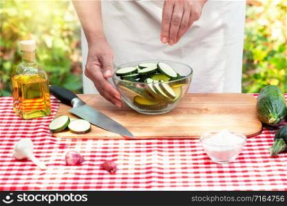 Female cook salt sliced zucchini in a transparent bowl. Checkered tablecloth on the table, natural green background. The concept of cooking natural eco-friendly dishes. Female cook salt sliced zucchini in a transparent bowl