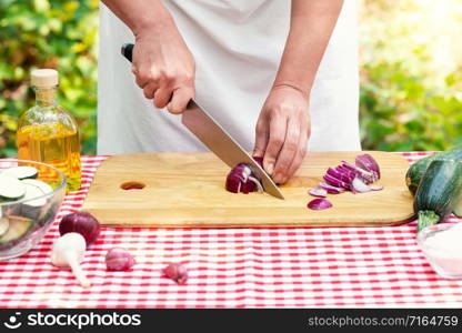 Female cook slices red onions. Checkered tablecloth on the table, natural green background. The concept of cooking natural eco-friendly dishes. Female cook slices red onions