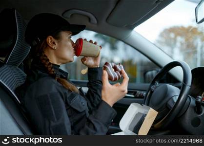 Female cop having lunch during workday. Woman police officer drinking coffee, eating donut in car. Female police officer having lunch during workday
