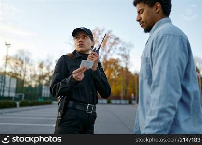 Female cop in black uniform checking male passerby ID document talking to walkie-talkie. Female cop checking male passerby ID document