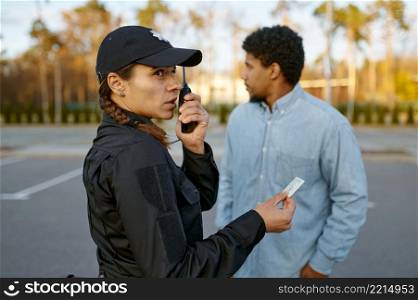 Female cop in black uniform checking male passerby ID document talking to walkie-talkie. Female cop checking male passerby ID document