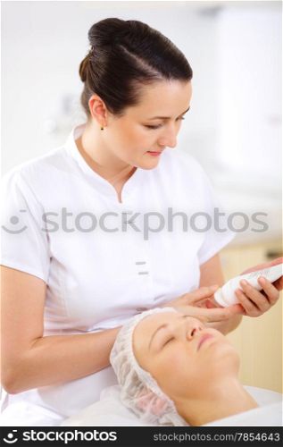 Female cosmetician looking at the tube with cosmetic while woman waiting for procedure starting