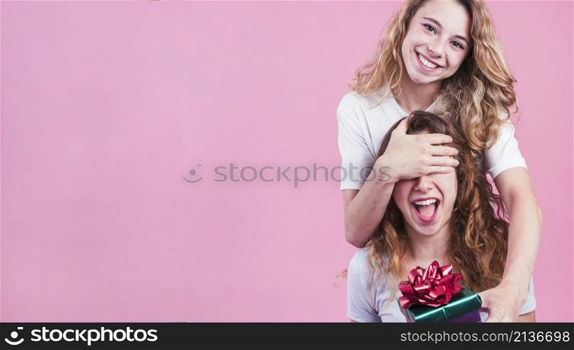 female covering her friend s eyes giving gift box against pink background