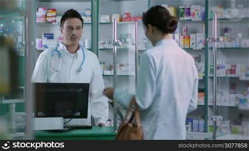Female customer giving prescription to pharmacist in pharmacy. Smiling male pharmacist with stethoscope in uniform reading prescription and checking availability in stock on computer while client waiting at the counter at local drugstore.