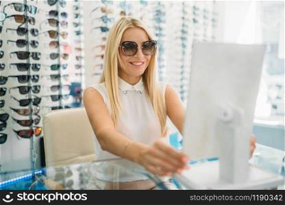 Female customer in sunglasses looks at the mirror in optics store. Selection of eyes protection in glasses shop, optometry concept