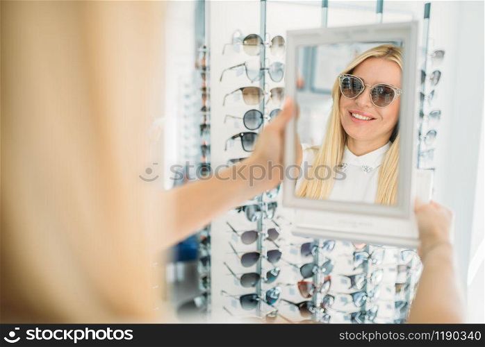 Female customer shooses sunglasses in optics store. Selection of eyes protection in glasses shop, optometry concept