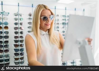 Female customer shooses sunglasses in optics store. Selection of eyes protection in glasses shop, optometry concept. Female customer shooses sunglasses in optics store
