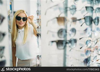 Female customer tries on sunglasses in optics store, showcase with spectacles on background. Selection of eyes protection in glasses shop, optometry concept