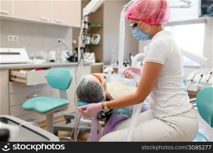 Female dentist holds mask, patient in chair, stomatology clinic. Doctor in uniform, medical worker, stomatologist, medicine and health, professional mouth care and hygiene, dentistry. Female dentist holds mask, patient in chair