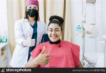 Female dentist with satisfied patient smiling at camera and giving thumb up, Female dentist with patient smiling and giving thumb up. Portrait of female dentist with patient smiling at camera in office