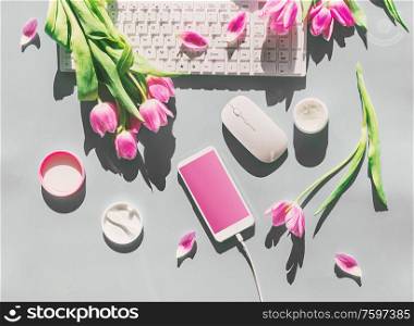 Female desktop with light pink tulips flowers bunch, cosmetics, PC keyboard , smartphone with blank screen and wire . Top view. Mothers day . Springtime. Greeting