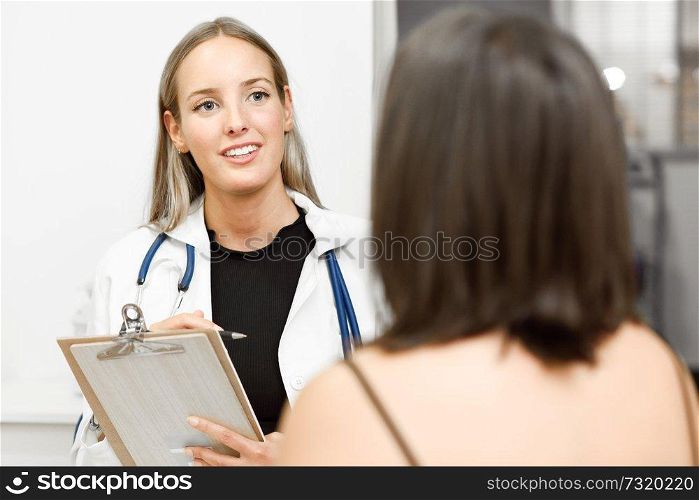 Female doctor explaining diagnosis to her patient. Brunette woman having consultation with smiling blonde girl in medical office.. Female doctor explaining diagnosis to her young woman patient.