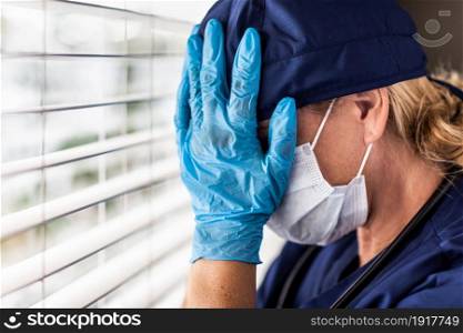 Female Doctor or Nurse With Head In Hands At Window Wearing Medical Face Mask and Surgical Gloves.
