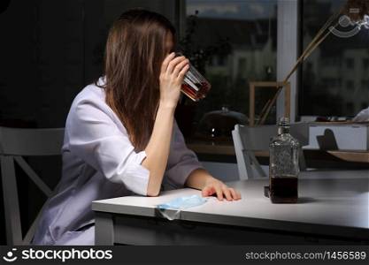 female doctor sitting at the table and hold glass with whiskey or cognac at home after hard work, depressed female drinking strong alcohol suffering from coronavirus problems. female doctor sitting at the table and hold glass with whiskey or cognac at home after hard work, depressed female drinking strong alcohol suffering from coronavirus problems.