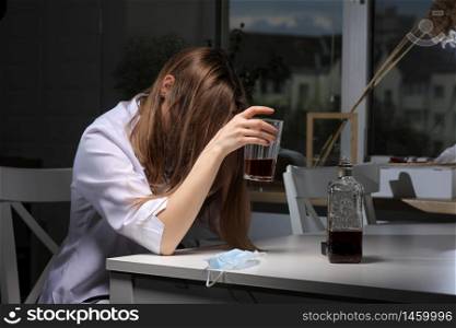 female doctor sitting at the table and hold glass with whiskey or cognac at home after hard work, depressed female drinking strong alcohol suffering from coronavirus problems.. female doctor sitting at the table and hold glass with whiskey or cognac at home after hard work, depressed female drinking strong alcohol suffering from coronavirus problems
