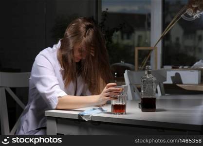 female doctor sitting at the table and hold glass with whiskey or cognac at home after hard work, depressed female drinking strong alcohol suffering from coronavirus problems. female doctor sitting at the table and hold glass with whiskey or cognac at home after hard work, depressed female drinking strong alcohol suffering from coronavirus problems.