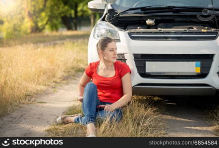 Female driver sitting on ground next to broken car and calling for assistance