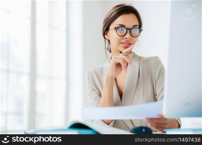 Female economist poses in coworking space, focused in monitor, verifies or checks figures from financial report, holds paper document and pen, has dark combed hair, wears formal business suit