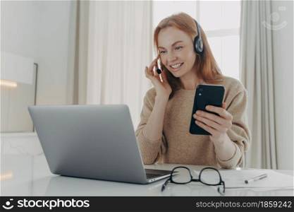 Female employee in headset has video call conference with partners holds modern smartphone watches webinar on laptop computer watches educational lecture poses at desktop against home interior. Female employee in headset has video call conference holds modern smartphone