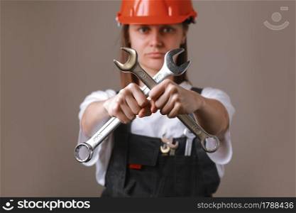 female engineer worker in a uniform and safety helmet with two cross wrenches in her hands and showing in camera. Equality between men and women. Repairwoman posing on a gray background. female engineer worker in a uniform and safety helmet with two cross wrenches in her hands and showing in camera. Equality between men and women. Repairwoman posing on a gray background.