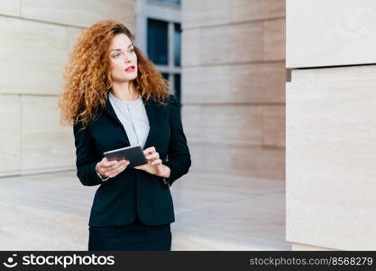 Female entrepreneur with curly bushy hair wearing formal clothes, holding modern tablet, looking with pensive expression aside, planning her working day. Woman developing project using touch pad