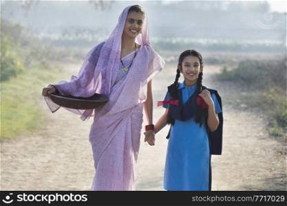 Female farmer or rural woman walking on village street along with her school going daughter carrying an iron gold pan.