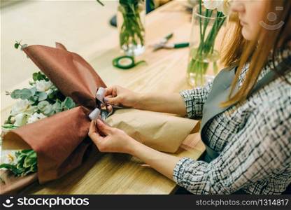Female florist attaches a bow to fresh flower composition. Floral business, bouquet preparation process, materials and tools for decoration on the table