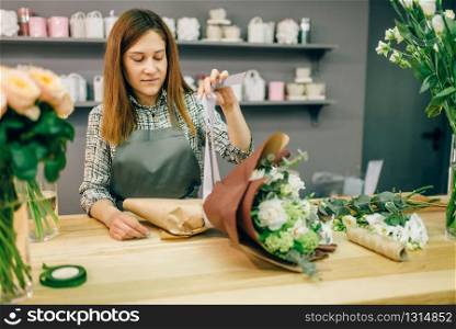 Female florist attaches a bow to fresh flower composition. Floral business, bouquet preparation process, materials and tools for decoration on the table. Florist attaches a bow to fresh flower composition