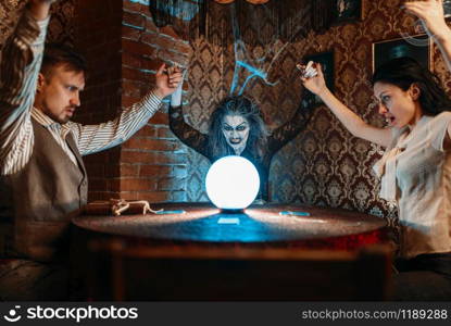 Female foreteller calls the spirits, witchcraft. Scary witch reads a magic spell over a crystal ball, young people hands up on spiritual seance