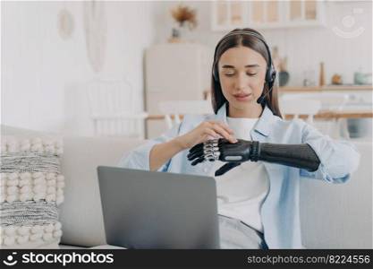 Female freelancer with disability wearing headset, working on laptop, setting bionic prosthetic arm before job. Distance education, elearning. Lifestyle of disabled people concept.. Girl with disability in headset customizes bionic prosthetic at laptop. Lifestyle of disabled people