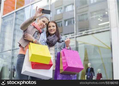 Female friends with shopping bags taking self portrait through mobile phone against store