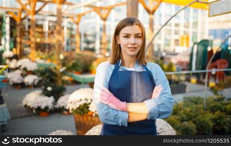 Female gardener poses in shop for gardening, saleswoman. Woman sells plants in florist store, seller in apron and gloves. Female gardener poses in shop for gardening