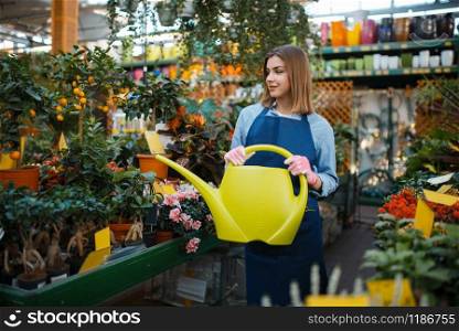 Female gardener with watering can takes care of plants in shop for gardening. Woman in apron sells flowers in florist store