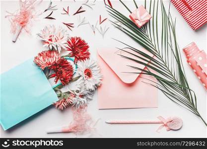Female greeting objects flat lay in coral and turquoise colors. Shopping bag with Flowers bunch, blank envelope with pen, palm leaves and wrapping paper on white background, top view.