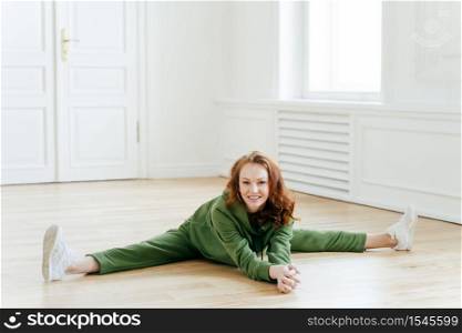 Female gymnast in sportswear, does leg split, sits on floor, poses in yoga room, has workout exercises, stretches legs, demonstrates splits, likes fitness. Active woman athlete poses indoor.