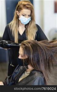 Female hairdresser drying her client&rsquo;s hair with a hairdryer, wearing protective masks in a beauty centre. Business and beauty concepts. Hairdresser drying her client&rsquo;s hair with a hairdryer wearing protective masks in a beauty centre.