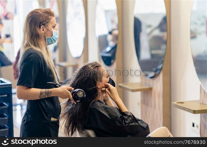 Female hairdresser drying her client’s hair with a hairdryer, wearing protective masks in a beauty centre. Business and beauty concepts. Hairdresser drying her client’s hair with a hairdryer wearing protective masks in a beauty centre.