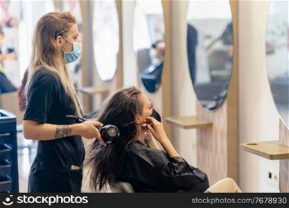 Female hairdresser drying her client’s hair with a hairdryer, wearing protective masks in a beauty centre. Business and beauty concepts. Hairdresser drying her client’s hair with a hairdryer wearing protective masks in a beauty centre.