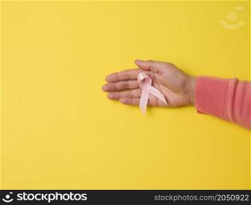 female hand hold a pink silk ribbon in the form of a loop on a yellow background. Symbol of the fight against breast cancer
