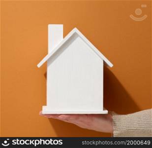 female hand hold a white miniature wooden house on a brown background. Real estate insurance concept, environmental protection, family happiness