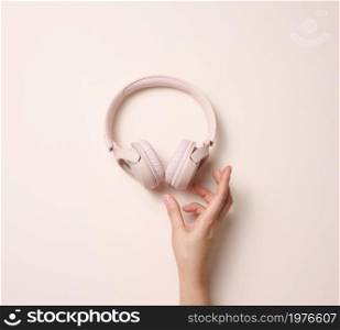 female hand hold pink wireless headphones on a beige background, top view