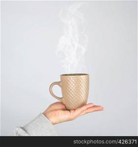 female hand holding a brown ceramic mug with a drink, thick steam rises from a cup, gray background